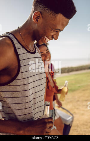 Smiling african man holding beer walking with friends outdoors on summer day. Friends having beers while walking to the picnic spot. Stock Photo