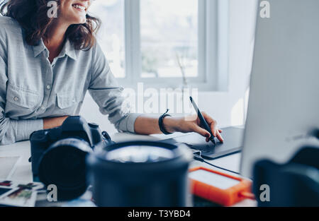 Female photographer working on computer using drawing pad at her desk. Young woman using graphic tablet and drawing pen to retouch the images. Stock Photo
