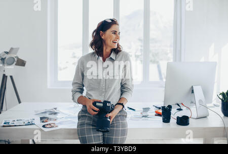 Cheerful woman photographer with a professional camera. Female in casuals leaning to the desk with digital camera in hand and looking away laughing. Stock Photo