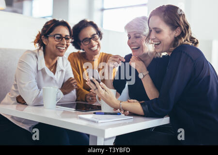 Happy women showing her phone to colleagues. Four businesswomen during break at office looking at mobile phone. Stock Photo