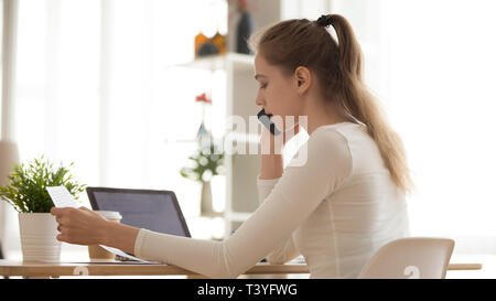Serious young woman talking on phone, reading paper document Stock Photo