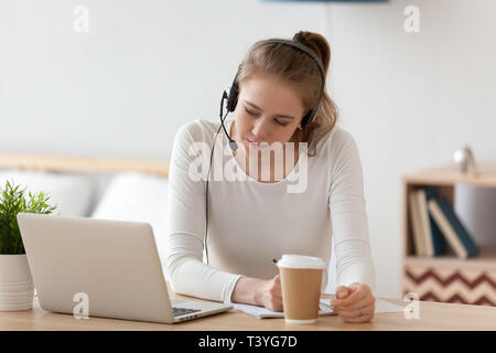 Focused woman in headset using laptop, writing notes Stock Photo