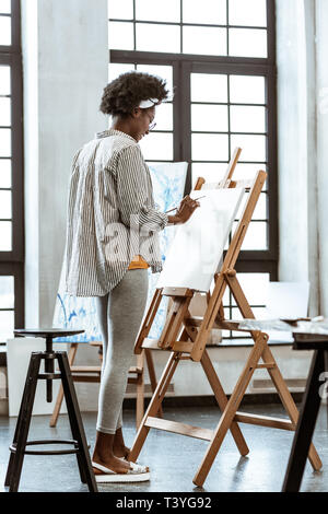 Truly inspired. Young curly artist feeling truly inspired while standing near drawing easel and painting Stock Photo