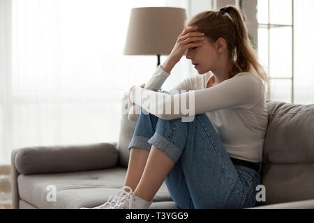 Upset woman frustrated by problem sitting on couch, embracing knees, Stock Photo