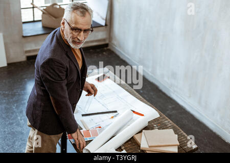 Standing near sketches. Grey-haired experienced interior designer wearing glasses standing near sketches Stock Photo