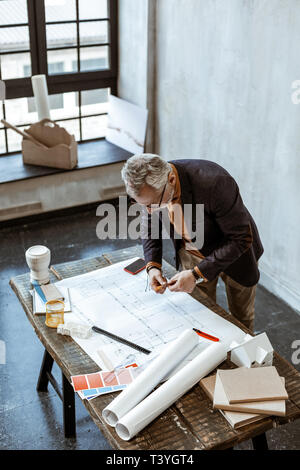 Making some notes. Grey-haired interior designer making some notes on the sketches in his light workshop Stock Photo