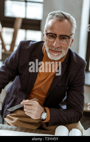 Holding the ruler. Prosperous dark-eyed interior designer holding the ruler while sitting at the table Stock Photo