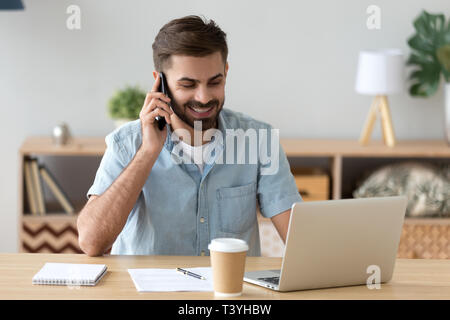 Smiling satisfied man talking on phone and using laptop at workplace Stock Photo