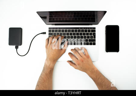 Hands of a businessman who is using his laptop computer and his smartphone on a white table. Modern workplace. External hard drive connected to the c Stock Photo
