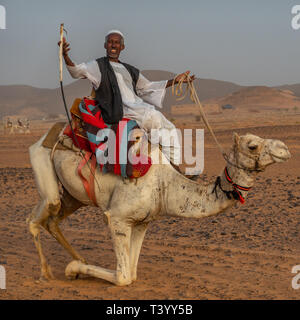 Meroe, Sudan, February 10., 2019: Older Sudanese man kneels down a camel for the pleasure of tourists Stock Photo