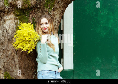 A young stylish woman is smiling, holding a present in her hand a bouquet of fresh mimosa flowers, March 8, Mother's Day. on a bright green background Stock Photo