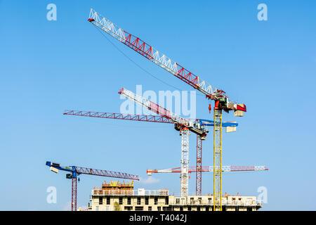 Five large tower cranes above a concrete building under construction against blue sky. Stock Photo