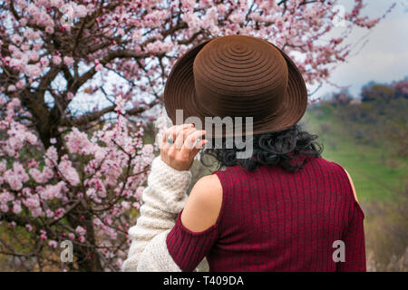 The girl in the hat on the background of the spring landscape with blooming trees. The girl in the burgundy dress and white sweater. View from the bac Stock Photo