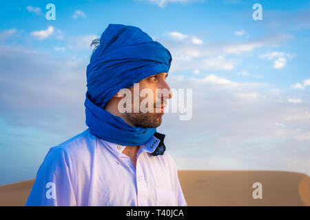 Full body of young Berber man wearing traditional Tuareg clothes walking on  sand dune while looking away in Merzouga desert Morocco stock photo