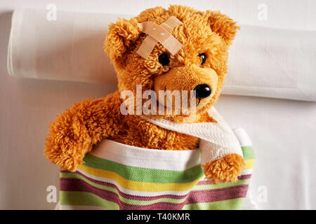 Cute little teddy bear with an arm in a sling and plaster or bandage on his head lying in bed under a colorful blanket in a concept of paediatric medi Stock Photo