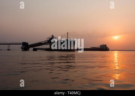 Port operations for managing and transporting iron ore at dawn and sunrise with 2 TSV ships - one at loading jetty, one waiting to dock. Sierra Leone Stock Photo