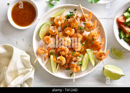 Delicious prawns skewers with greens,spices, lime and sauce on white plate over wooden table. Grilled shrimp skewers. Tasty seafood. Top view, flat la Stock Photo