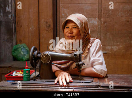 Friendly smiling woman with a pink headkerchief sitting behind an old fashioned sewing machine. Borobudur Village, Java, Indonesia Stock Photo