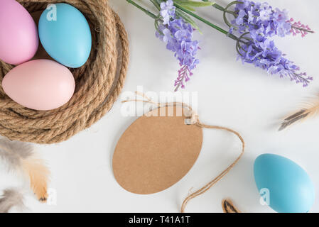 Blank rustic egg-shaped gift tag on white background with nest, pastel Easter eggs, feathers, and flowers Stock Photo
