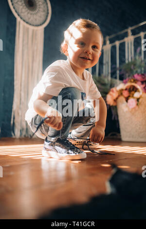 Toddler boy learns to tie the shoelaces on his sneakers opposite the window. Stock Photo