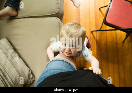 Whining blonde toddler boy clings to mother's leg while looking up Stock Photo