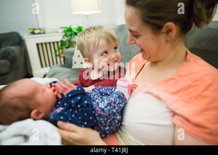 Happy blonde toddler boy looks up at mother holding newborn baby girl Stock Photo