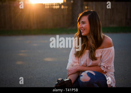 a beautiful smiling woman sits at sunset with her camera on a street Stock Photo