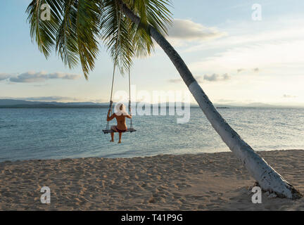 Rear view of woman in a beach swing attached to a palm tree in San Blas Islands. RF Travel destination, lifestyle / holiday concept. Panama, Oct 2018 Stock Photo