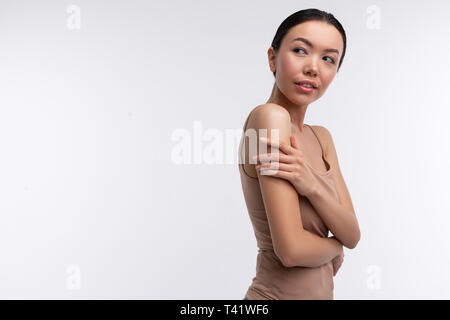 Skinny woman. Skinny dark-eyed woman wearing beige camisole standing near background touching her shoulder Stock Photo