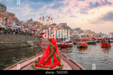 Young Indian girl in traditional dress (saree) on a wooden boat overlooking the historic Varanasi city architecture and Ganges river ghat at twilight. Stock Photo