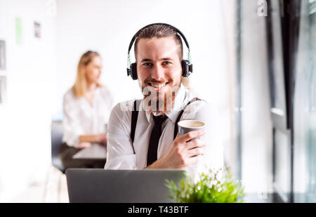 A portrait of young businessman with headphones and coffee in an office. Stock Photo