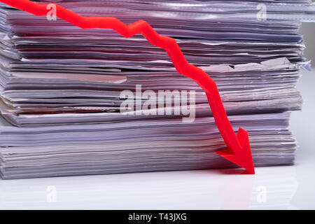 Red Diminishing Arrow Over White Desk With Background Of Stacked Paper Stock Photo