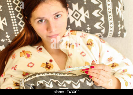 Top down view, blurred young woman laying in bed in her pyjama, holding thermometer (in focus). Grip / flu sickness concept.