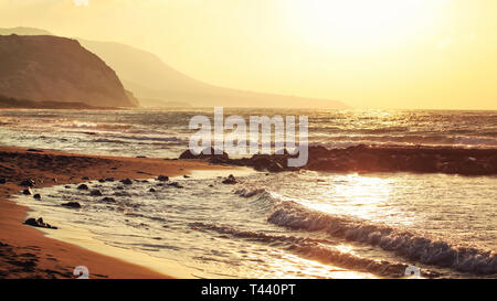 Empty wild beach lit by golden sunset. Small waves over rock, hills in hazy background. Stock Photo