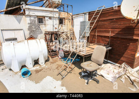Midday sun shining on garbage and discarded stuff (rusty desks and old chair) partly covered in sand, behind beach restaurant. Akdeniz, Northern Cypru Stock Photo