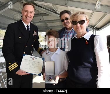 Panama City, FL -- Naval Support Activity Panama City commanding officer Cmdr. Jay Sego presents Kristopher Parker, grandson of World War II veteran Gunner's Mate 2nd Class Wheeler R. Ervin, the Purple Heart during a ceremony Mar. 14, 2019. Also present to receive the medal was son-in-law Terry Parker and daughter Nicole Ervin Parker.  In addition, Ervin's family also received the Navy-Marine Corps Commendation Medal with Valor for his actions aboard the Liberty Ship SS William W. Gerhard in August 1943.  As a member of the Navy's Armed Guards whose mission was to protect the Liberty Ship conv Stock Photo