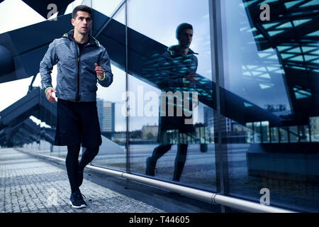 Sports man doing his run workut in scenery of modern city architecture - glass wall of building. Stylish sportswear. Stock Photo