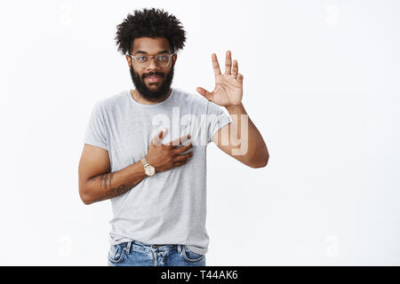 You have my word. Portrait of charming confident, smiling african american man giving promise raising hand and holding arm on heart as giving oath or  Stock Photo