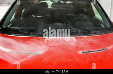 Front hood of red car washed in self service carwash, white water spraying on metal, blurred seats and interior background Stock Photo