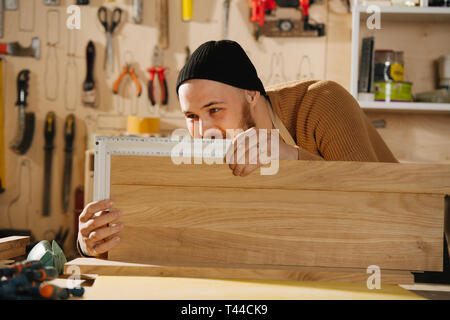 Portrait of working handsome middle aged bearded carpenter in a watch cap. He is making furniture on order in a workshop. Checking flatness of two sur Stock Photo