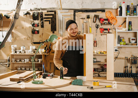 Portrait of working handsome middle aged bearded carpenter in a watch cap. He is making furniture on order in a workshop. Taking few seconds between t Stock Photo