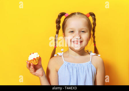 Closeup portrait of a cheerful little sweet tooth girl on a yellow background. The child smeared his nose with cream and holding a cake in his hands. Stock Photo