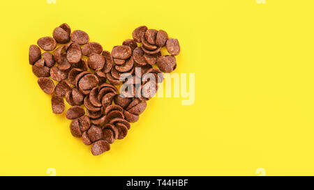 Top down view, chocolate cornflakes at yellow board arranged in shape of heart, space for text on right side Stock Photo