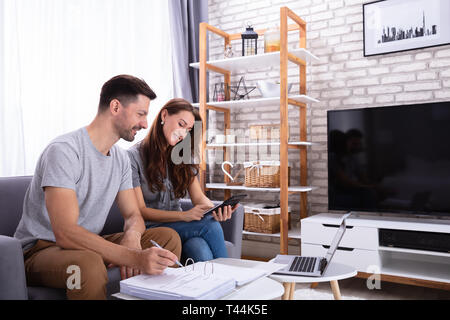 Smiling Young Couple Sitting On Sofa Calculating Invoice At Home Stock Photo