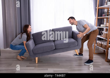 Side View Of A Smiling Young Couple Lifting Sofa In Living Room Stock Photo