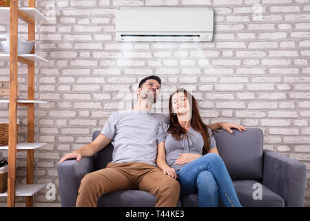 Happy Young Couple Sitting Under Air Conditioner On Couch At Home Stock Photo