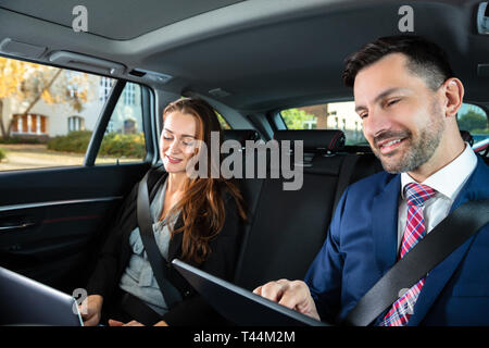 Two Happy Businesspeople Using Electronic Devices While Traveling By Car Stock Photo