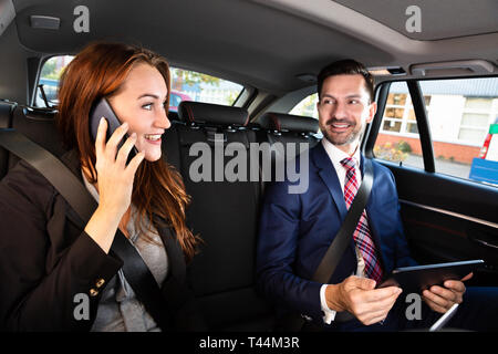 Happy Young Businesswoman Sitting Inside Car Beside Her Male Colleague Using Smartphone Stock Photo