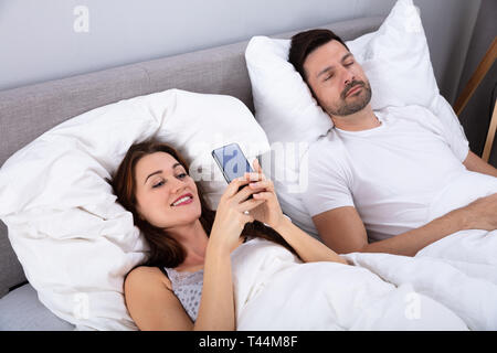 Smiling Woman Using Smartphone While Her Husband Sleeping On Bed In Bedroom Stock Photo