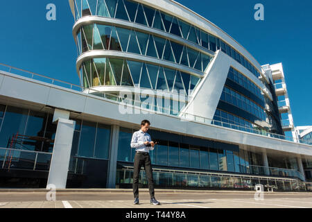 young businessman in glasses with bag in hands stands near the building. freelancer holds a tablet and works on the street Stock Photo